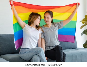 Millennial Asian Young Happy Female Lesbian LGBTQ Lover Couple Sitting Smiling Together On Cozy Sofa Holding Big Rainbow Gay Pride Unity Flag Showing Same Sex Love Equality In Living Room At Home.