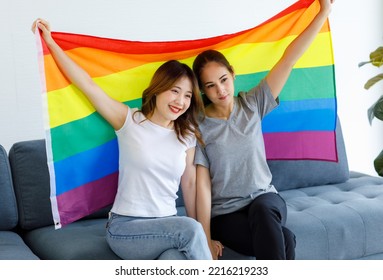 Millennial Asian Young Happy Female Lesbian LGBTQ Lover Couple Sitting Smiling Together On Cozy Sofa Holding Big Rainbow Gay Pride Unity Flag Showing Same Sex Love Equality In Living Room At Home.