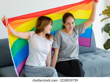 Millennial Asian Young Happy Female Lesbian LGBTQ Lover Couple Sitting Smiling Together On Cozy Sofa Holding Big Rainbow Gay Pride Unity Flag Showing Same Sex Love Equality In Living Room At Home.