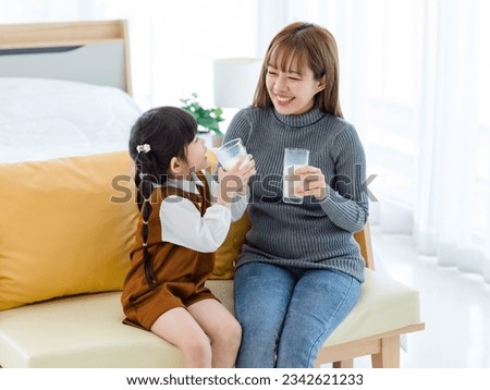 Millennial Asian young pretty female teenager mother nanny babysitter in casual outfit sitting on sofa smiling holding serving delicious milk glass to little cute preschooler daughter girl drinking.