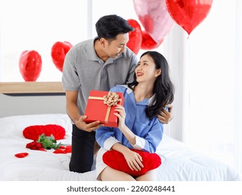Millennial Asian young romantic lover couple male boyfriend giving present wrapped gift box surprised to female girlfriend on bed decorated with heart shape balloon on valentine day anniversary.