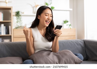 Millennial asian young woman looking mobile phone laughing with good news or discount voucher for shopping online at home.Happy and cheerful woman looking on cellphone app read message feel excited - Shutterstock ID 2107699415
