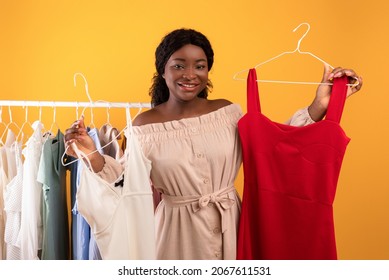 Millennial black female choosing between two trendy dresses on orange studio background. Young African American woman shopping for new clothes, selecting stylish wardrobe