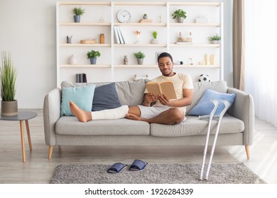 Millennial Black Guy With Plastered Leg Sitting On Sofa, Reading Book At Home, Pair Of Crutches Standing Nearby. Young African American Man Having Broken Limb, Resting On Couch With Interesting Story