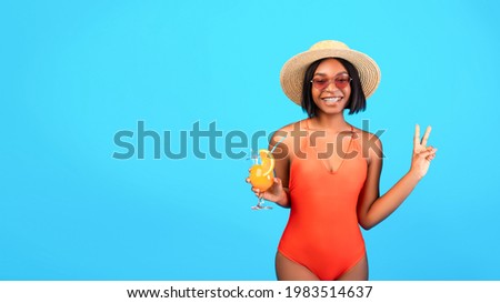 Millennial black lady in straw hat and bikini holding exotic cocktail and showing peace gesture on blue studio background, panorama with copy space. Young woman with cool drink having beach party