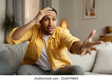Millennial black man emotionally reacting to shocking news or defeat of sports team on television, sitting on couch at home. Young African American guy expressing disbelief or indignation indoors