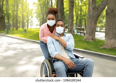 Millennial black woman hugging her handicapped boyfriend in wheelchair, wearing face masks, going for walk at city park. African American impaired couple spending time outdoors during coronavirus