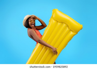 Millennial black woman in swimsuit holding yellow inflatable lilo on blue studio background. Young African American lady with pool float having tropical summer vacation or beach party