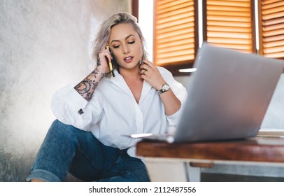 Millennial blonde woman with digital netbook technology using wireless connection on mobile phone for calling and talking indoors, skilled freelancer making consultancy conversation via cellphone