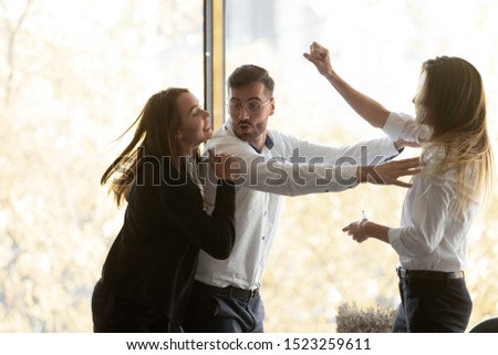 Millennial businessman calming down, setting apart two aggressive fighting female colleagues at workplace. Young women quarrelling, having conflict or misunderstanding during workday at office.