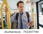 Millennial businessman is commuting on a tram in Melbourne, Victoria. He is watching something on his smart phone with headphones while standing and holding on to the rail. 