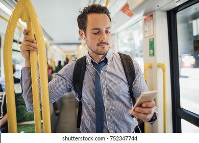 Millennial businessman is commuting on a tram in Melbourne, Victoria. He is watching something on his smart phone with headphones while standing and holding on to the rail. 