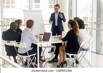 Millennial businessman in suit stand hold meeting make flip chart presentation for diverse colleagues, male coach or speaker talk present project graph on whiteboard training multiethnic work team - Shutterstock ID 1389051566