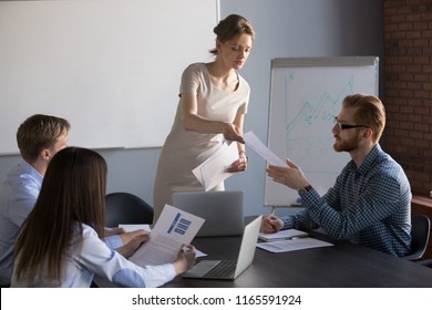 Millennial Businesswoman Give Handout Materials To Work Team Members During Flipchart Presentation, Female Speaker Or Coach Share Documents To Workers, Presenter Hand Papers To Consider Or Analyze