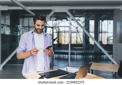 Millennial Caucasian Businessman Holding Business Card And Watching Something On Mobile Phone. Concept Of Modern Successful Man. Young Smiling Guy In Wireless Earphones At Desk In Open Space Office