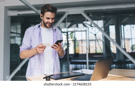 Millennial Caucasian Businessman Holding Business Card And Watching Something On Smartphone. Concept Of Modern Successful Man. Young Smiling Guy In Wireless Earphones At Desk In Open Space Office