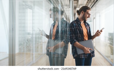 Millennial caucasian businessman with laptop using and watching on smartphone during work. Concept of modern successful man. Young bearded smiling male entrepreneur in glasses in open space office - Powered by Shutterstock