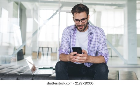 Millennial Caucasian Businessman Typing And Watching On Mobile Phone. Concept Of Modern Successful Man. Young Focused Guy In Glasses Sitting On Leather Bench With Opened Laptop In Open Space Office