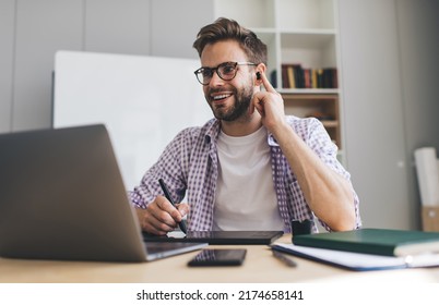 Millennial caucasian businessman working on laptop, writing in graphics tablet and correcting earphone in ear. Concept of modern successful man. Young smiling guy in glasses at desk in office - Powered by Shutterstock