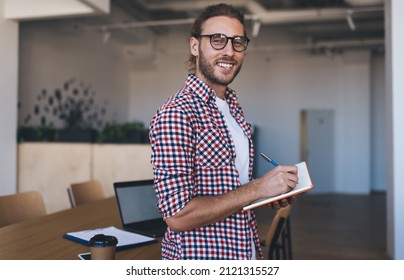 Millennial caucasian businessman writing in notebook and looking at camera. Concept of modern successful man. Young smiling male entrepreneur in glasses stand and work at table in open space office - Powered by Shutterstock