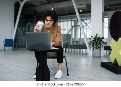 Millennial Caucasian Businesswoman Drinking Coffee And Using Laptop During Work. Concept Of Modern Successful Woman. Young Focused Girl In Glasses Sitting In Armchair At Table In Open Space Office