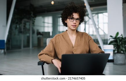 Millennial Caucasian Businesswoman Looking At Camera During Working On Laptop. Concept Of Modern Successful Woman. Young Serious Curly Girl In Glasses Sitting In Armchair In Open Space Office