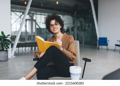 Millennial Caucasian Businesswoman Looking At Camera During Writing In Notebook At Work. Concept Of Modern Successful Woman. Young Smiling Girl In Glasses Sitting In Armchair At Open Space Office