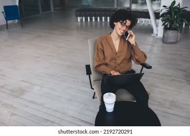 Millennial Caucasian Businesswoman Talk On Smartphone And Watch On Laptop During Work. Concept Of Modern Successful Woman. Young Smiling Curly Girl In Glasses Sitting In Armchair At Table In Office