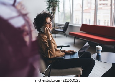 Millennial Caucasian Businesswoman Talking On Smartphone During Working On Laptop. Concept Of Modern Successful Woman. Young Smiling Girl In Glasses Sitting In Armchair At Table In Open Space Office