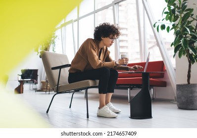 Millennial Caucasian Businesswoman Typing On Laptop During Work. Concept Of Modern Successful Woman. Young Curly Girl In Glasses With Coffee Sitting In Armchair At Table In Open Space Office