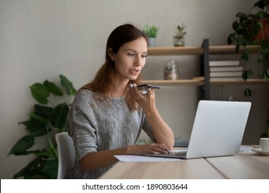 Millennial Caucasian female employee work on laptop from home office record audio message on cellphone. Woman talk activate digital voice assistant on smartphone, consult client online on computer. - Powered by Shutterstock