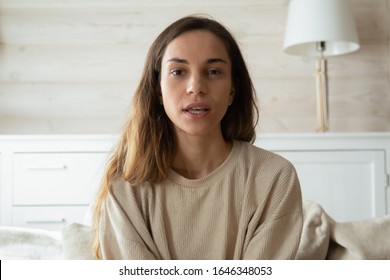Millennial Caucasian girl sit on couch in living room look at camera having webcam conversation, young woman talk speak on video call using wireless internet connection at home, female blogger record