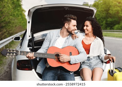 Millennial Caucasian man and his girlfriend traveling by auto, sitting in car trunk with suitcase and guitar, creating music. Young loving couple having road trip on summer holidays - Powered by Shutterstock