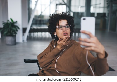 Millennial Caucasian Woman Blowing Kiss During Taking Selfie On Smartphone And Listening Music In Earphones. Concept Of Modern Successful Woman Lifestyle. Young Girl In Glasses Sitting In Armchair