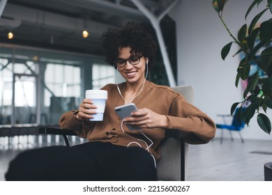 Millennial Caucasian Woman Drinking Coffee, Listening Music In Earphones And Watching On Smartphone. Concept Of Modern Successful Woman Lifestyle. Young Smiling Girl In Glasses Sitting In Armchair