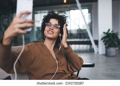 Millennial Caucasian Woman Gesticulating During Taking Selfie On Smartphone And Listening Music In Earphones. Modern Successful Woman Lifestyle. Young Smiling Girl In Glasses Sitting In Armchair