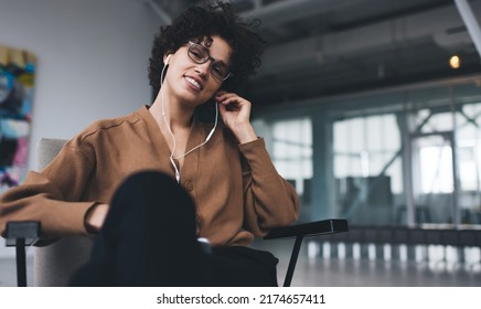 Millennial Caucasian Woman Listening Music In Earphones And Looking At Camera. Concept Of Modern Successful Woman Lifestyle. Young Smiling Girl In Glasses Sitting In Armchair At Open Space Office