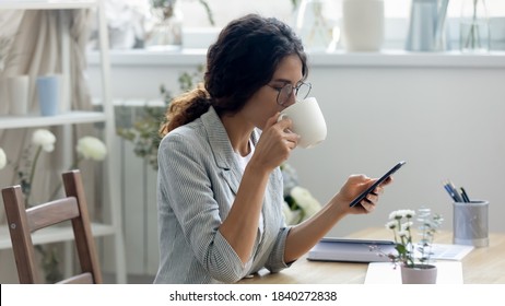 Millennial Caucasian woman sit at desk in home office browse surf internet on smartphone drink coffee enjoy morning at workplace. Young female text message on cellphone gadget. Communication concept. - Powered by Shutterstock