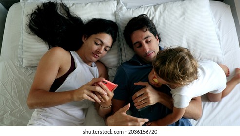 Millennial Couple In Bed With Infant Toddler Boy Wanting Attention. Man And Woman Lying Down Looking At Cellphone, Top View