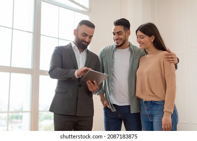 Millennial Couple Buying New Apartment, Standing In Empty Room, Professional Real Estate Agent In Suit Showing Digital Tablet With Build Project, Discussing House Plan, Selling Flat To Happy Family