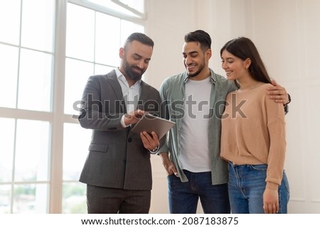Millennial Couple Buying New Apartment, Standing In Empty Room, Professional Real Estate Agent In Suit Showing Digital Tablet With Build Project, Discussing House Plan, Selling Flat To Happy Family