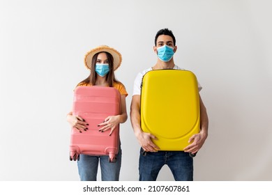 Millennial couple in face masks holding their suitcases in hands, standing over light wall. Vaccinated man and woman ready for vacation, enjoying post-quarantine travels - Powered by Shutterstock