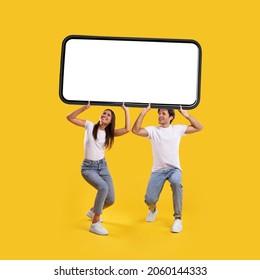 Millennial couple holding huge heavy smartphone with white empty screen on yellow orange studio background. Mock up for mobile app or new website advertising, great offer. Cellphone display template
