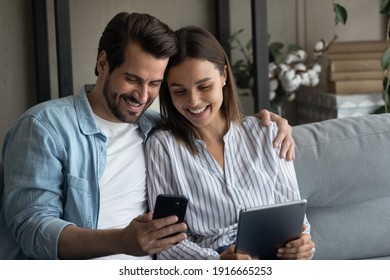 Millennial Couple In Love Confident Internet Users Sit On Couch Hold Phone Tablet Exchange Data Share Information Between Devices. Young Husband Wife Send Receive Files App From Cell To Pad Using Wifi