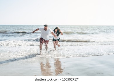 A Millennial Couple Running And Splashing Water On The Beach In California. Wearing Summer Clothing Getting Wet. Both Are Smiling And Having Fun. The Male Almost Falls.