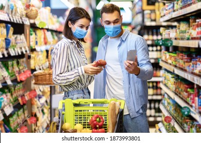 Millennial Couple Using Smartphone Scanning Food Product In Supermarket Doing Grocery Shopping Together And Buying Supplies. Buyers Wearing Face Masks Reading Checklist On Phone Standing In Store