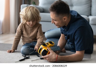 Millennial dad and cute preschooler son engaged in motor mechanic games on heating floor, repairing toy plastic car with screwdrivers. Father and kid playing together, enjoying activities at home