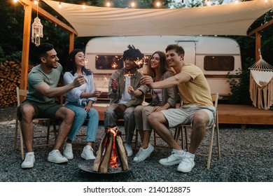 Millennial diverse friends sitting near bonfire with lit sparklers, having fun, celebrating something together on camping trip, traveling in motorhome, enjoying friendly conversation outdoors - Powered by Shutterstock