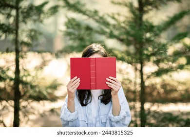 A millennial european person is holding a red hardcover book in front of their face, standing among a grove of trees that diffusely illuminate the scene with dappled sunlight
