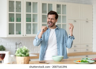 Millennial excited man cooking healthy tasty vegetarian breakfast in kitchen, holds whisk singing having fun, entertaining enjoy good morning new day and favourite pastime. Talent, home hobby concept